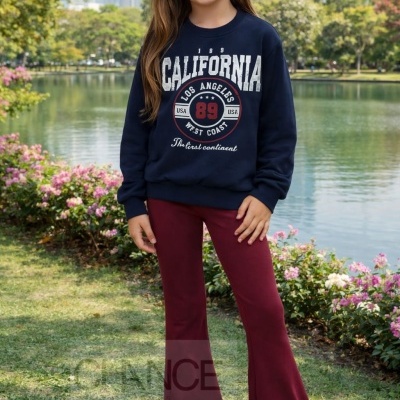 Menina com sweatshirt azul escuro com texto CALIFORNIA e calças bordeaux em parque junto a lago.