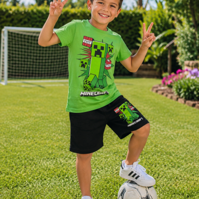 Menino com camisola verde e calções pretos estampados de Minecraft, com um pé em bola de futebol num campo gramado