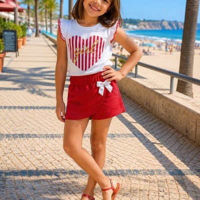 Menina vestindo camiseta branca com padrão de coração, calção vermelho e sandálias vermelhas com flores em passeio perto da praia.