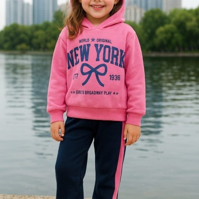 Menina sorridente com hoodie rosa e calças azul-escuras junto a um lago em ambiente urbano