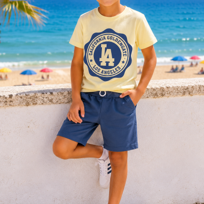 Rapaz jovem com t-shirt amarela e calções azuis junto ao mar numa praia com guarda-sóis.
