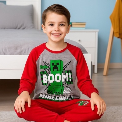 Menino sentado no chão a usar pijama de Minecraft cinza e vermelho com uma cama branca e parede azul ao fundo.