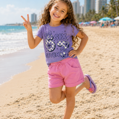 Menina na praia com camisola lilás estampada, calções cor-de-rosa e sandálias lilás.