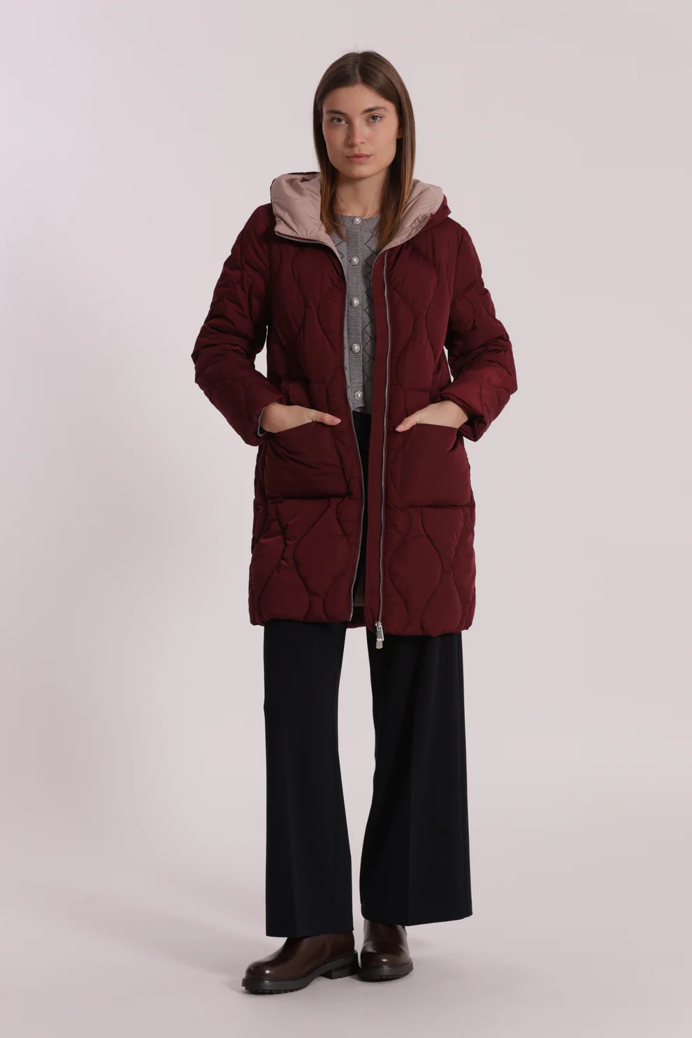 Casaco acolchoado bordeaux com capuz e bolsos, usado por mulher com calças pretas e botas castanhas.