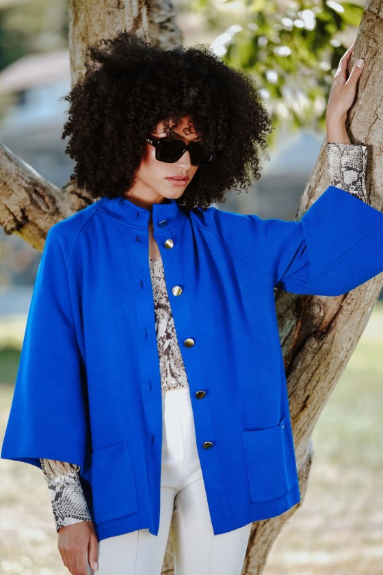 Mulher com casaco azul royal, blusa animal print, calças brancas e óculos escuros junto a uma árvore