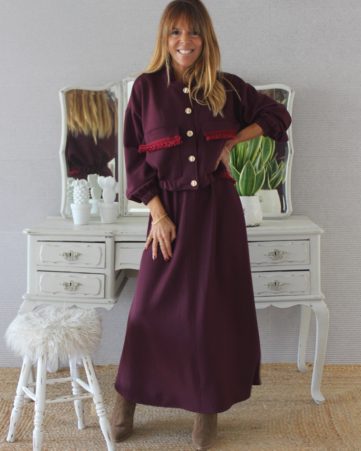 mulher com conjunto vinho de casaco com botões e saia comprida ao pé de móvel branco com planta e banco felpudo
