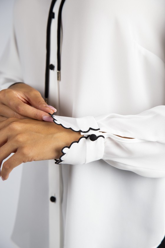 Blusa branca com botões pretos e punhos ondulados com remate preto.