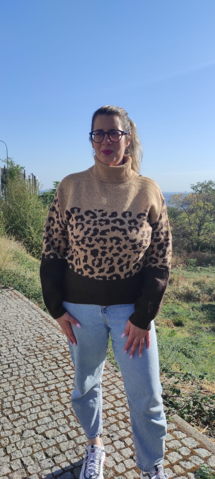 Mulher com suéter de malha com padrão leopardo, jeans e tênis brancos num caminho de pedra