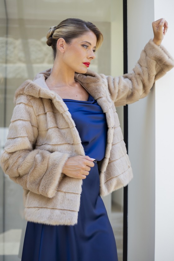 Mulher com casaco bege de pelúcia e vestido azul junto a uma parede branca