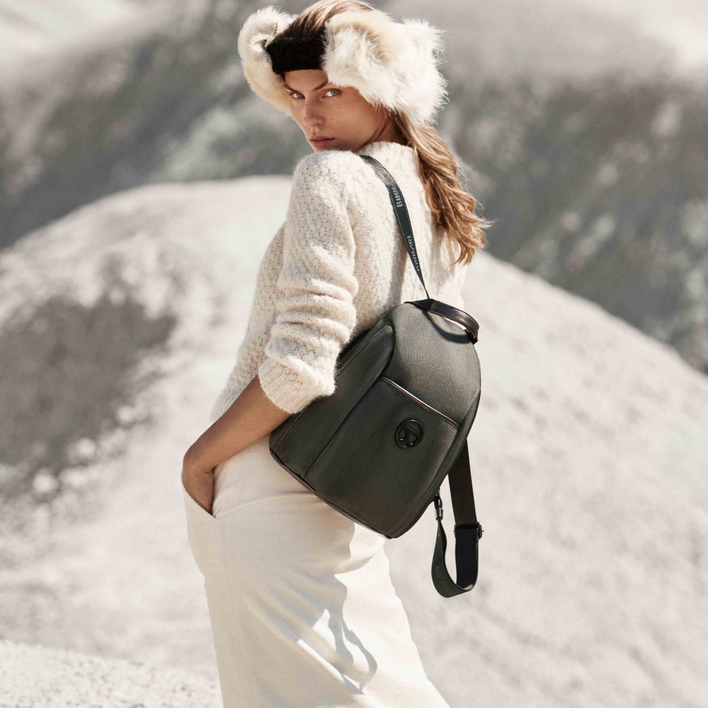 Mulher com mochila preta texturizada e alça com texto branco em cenário de montanhas.