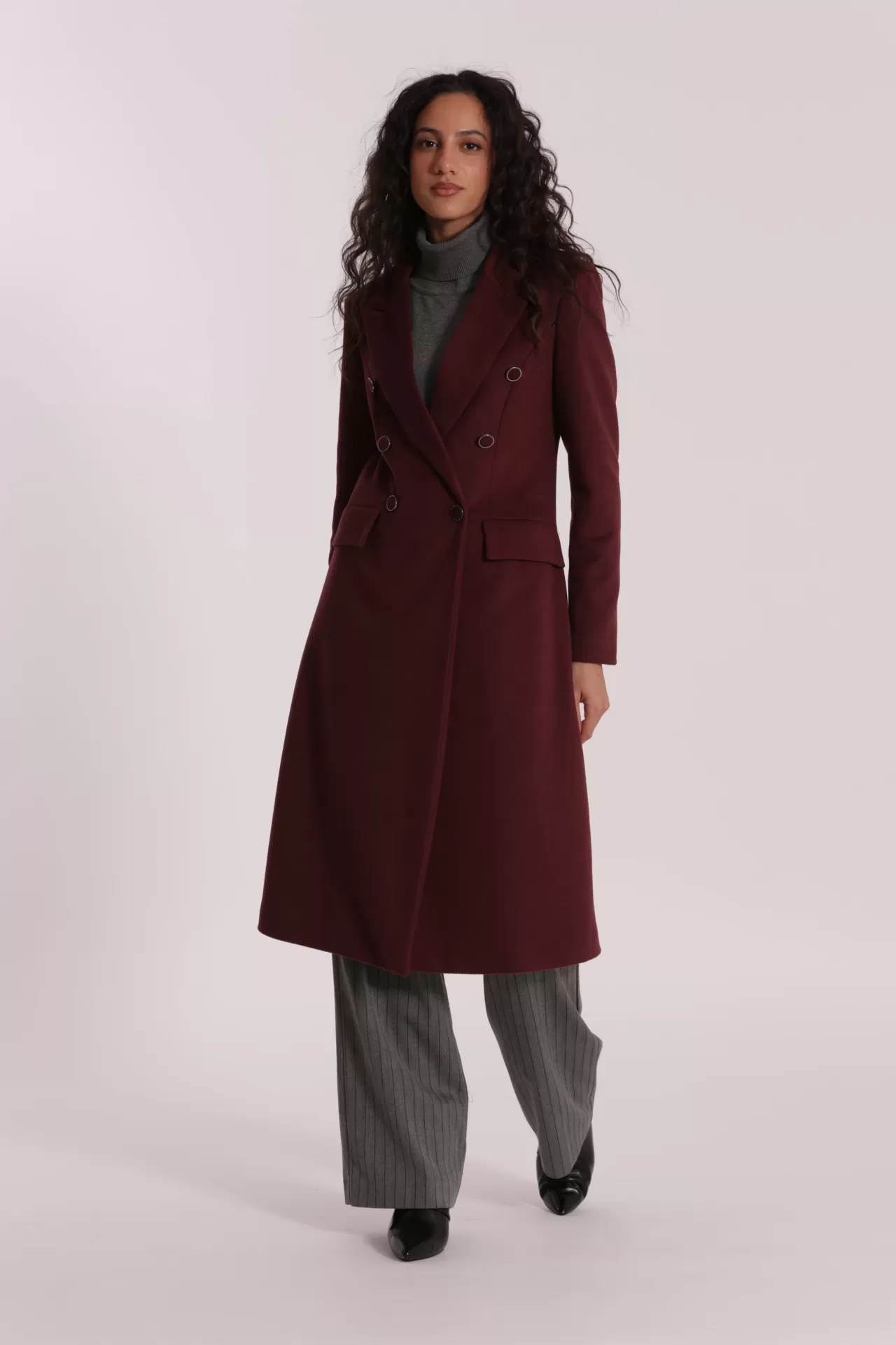 Mulher a usar casaco comprido bordô com botões, camisola cinzenta, calças cinzentas de riscas e sapatos pretos.