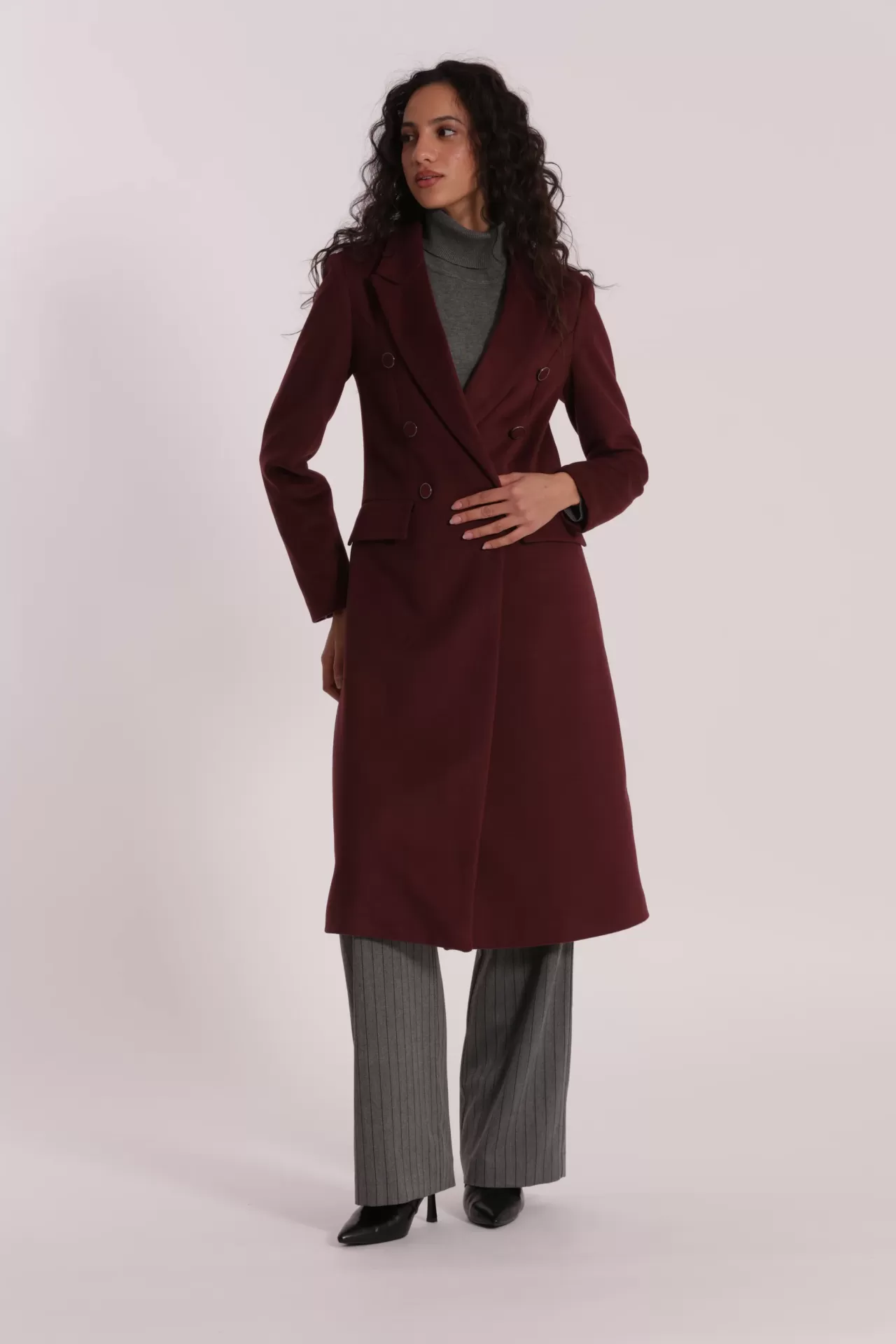 Mulher veste casaco longo bordô, blusa cinza e calças cinza riscas com sapatos pretos