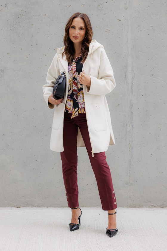 Mulher com casaco branco, blusa estampada, calças vinho e sapatos pretos em fundo cinzento.