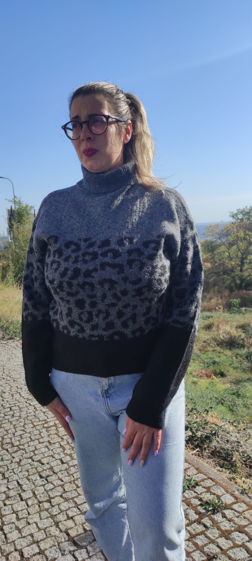 Mulher de camisola cinzenta com padrão animal print preto e calças azuis na rua com vegetação ao fundo
