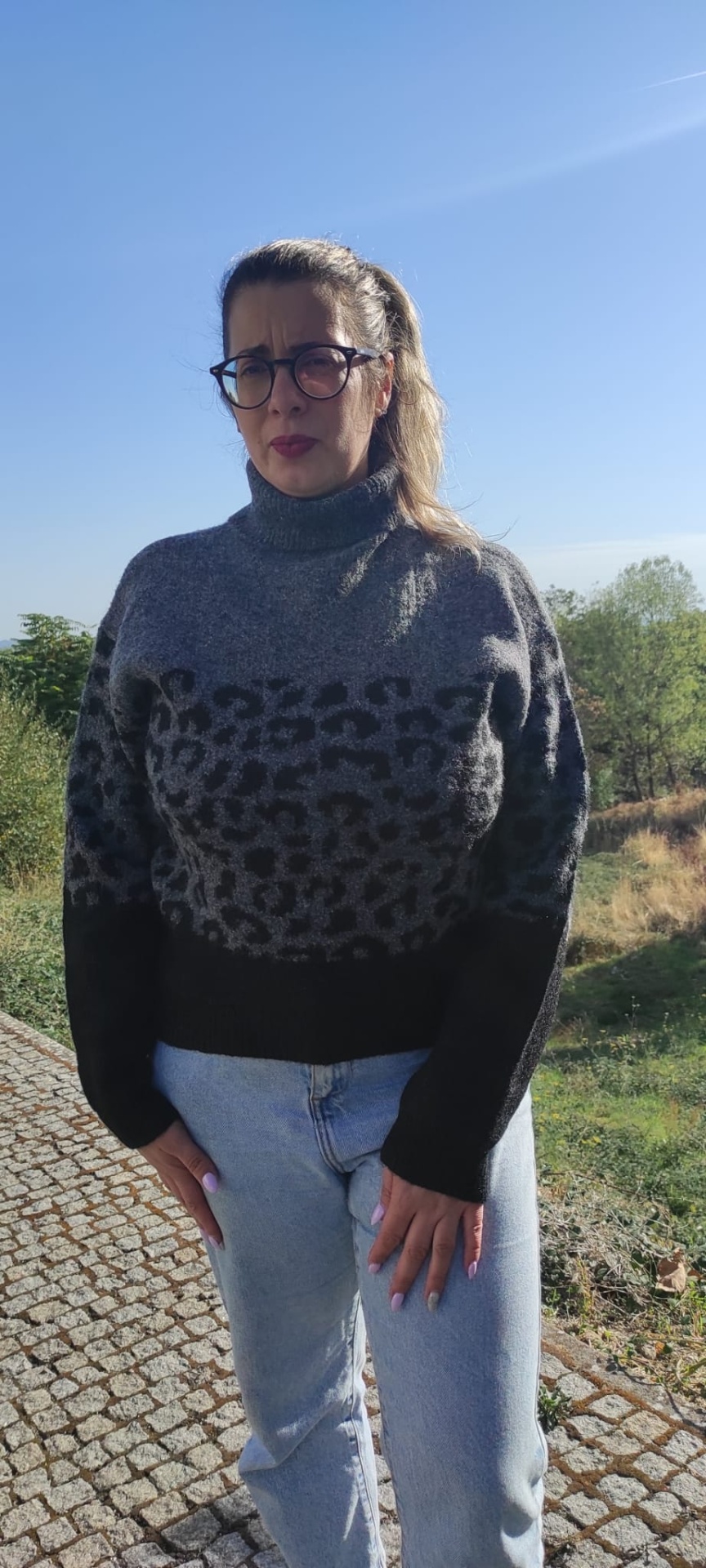 Mulher com casaco de malha cinzento com padrão preto, calças de ganga e óculos, ao ar livre com céu azul