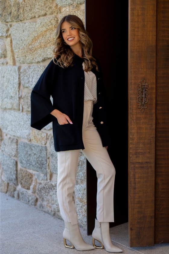 Mulher vestindo casaco preto, blusa branca com bolinhas, calças bege e botas bege junto a parede de pedra e porta de madeira