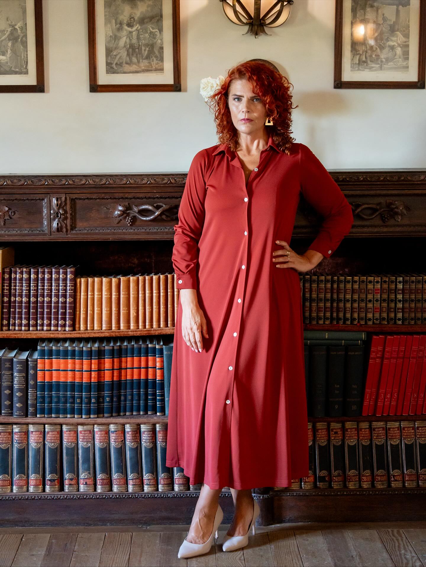 Vestido longo vermelho com botões e sapatos brancos de salto em biblioteca com estante de livros.