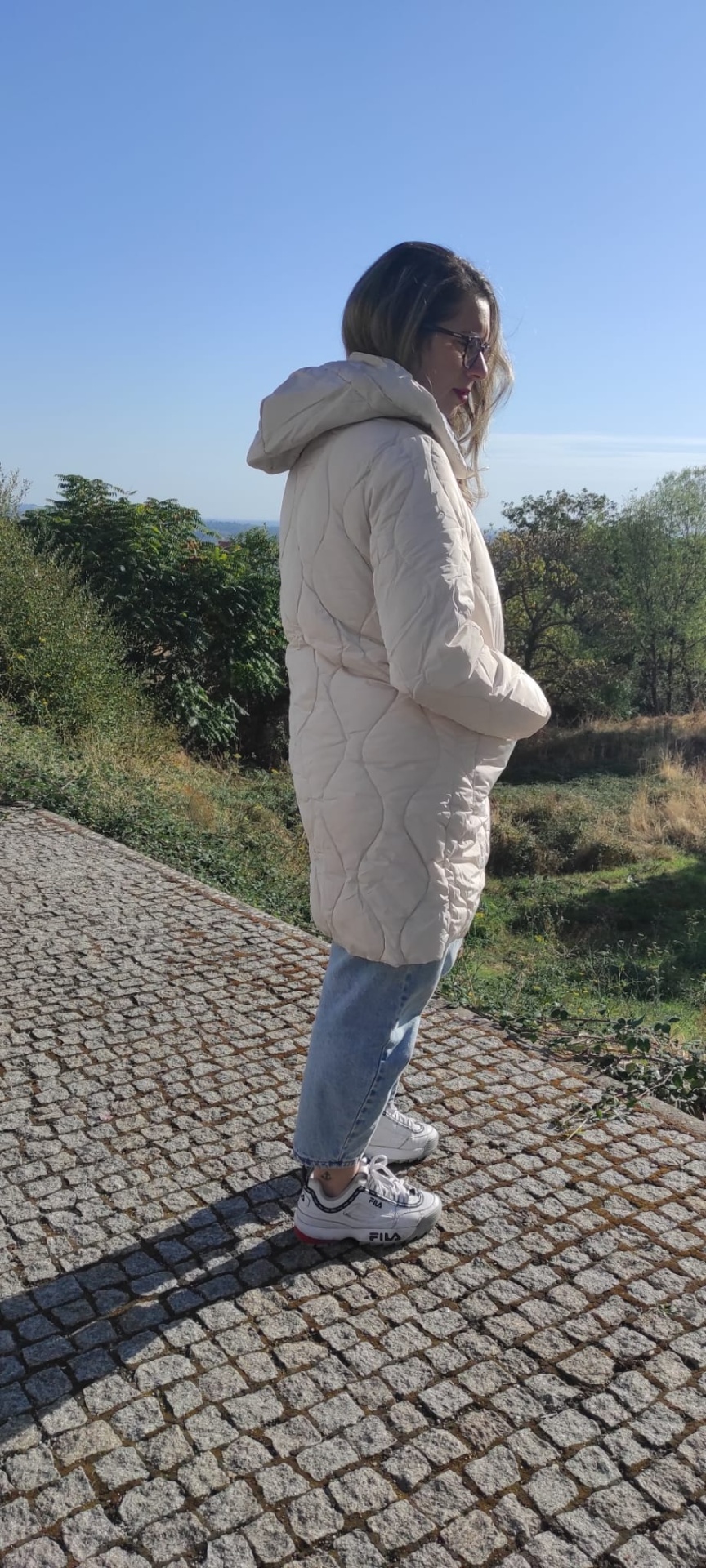 Mulher de perfil com casaco acolchoado bege, calças de ganga e ténis brancos FILA num passeio de pedra ao ar livre