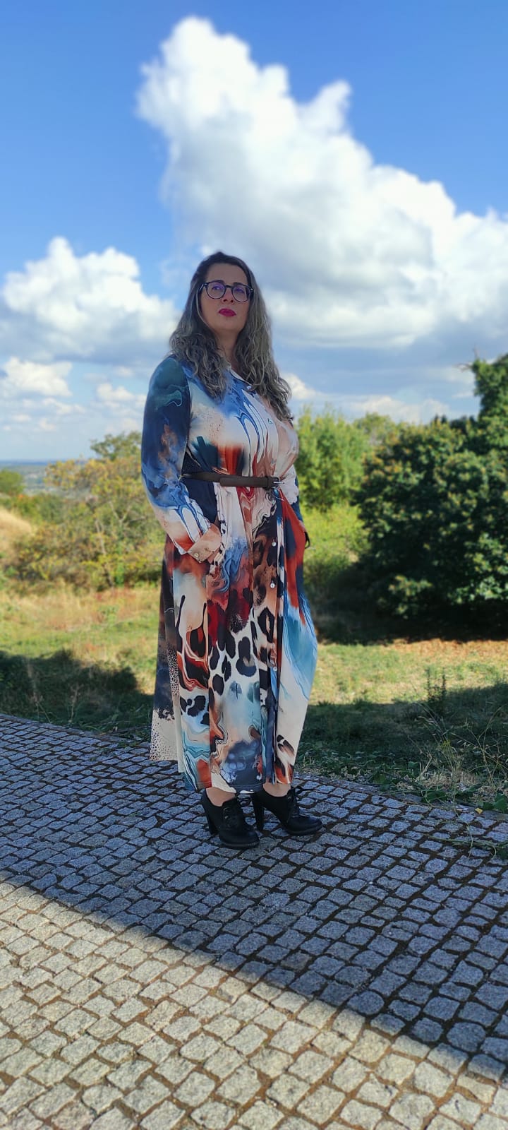 Mulher com vestido comprido colorido com padrão abstrato e cinto castanho, sapatos pretos, em exterior com calçada e vegetação.