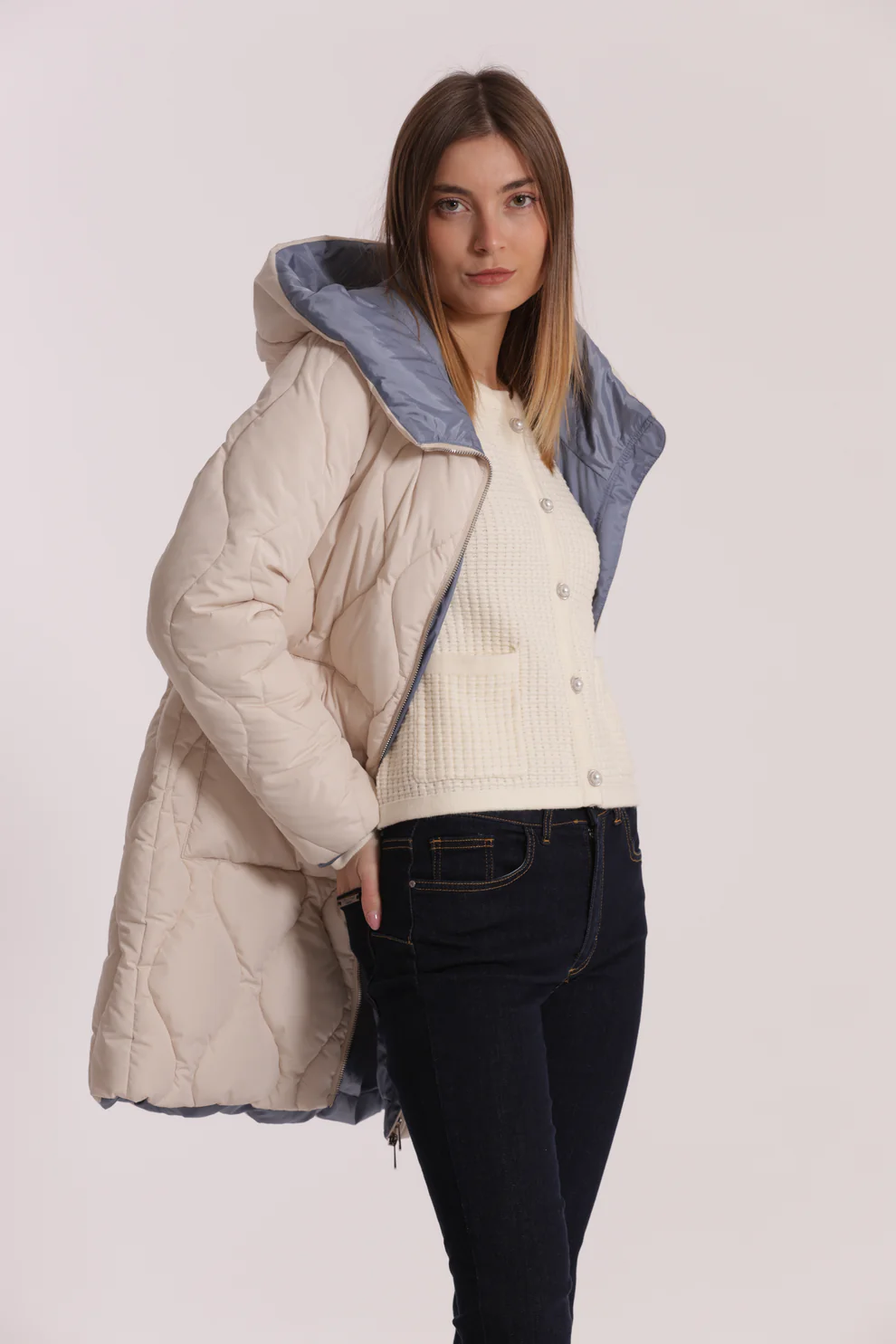Mulher com casaco acolchoado bege e capuz azul cinzento, camisa creme e calças de ganga escuras.