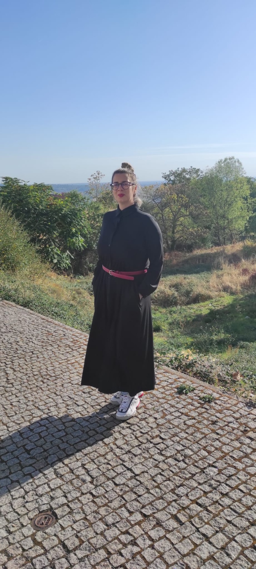 Mulher com vestido comprido preto e cinto rosa em caminho de calçada de pedra