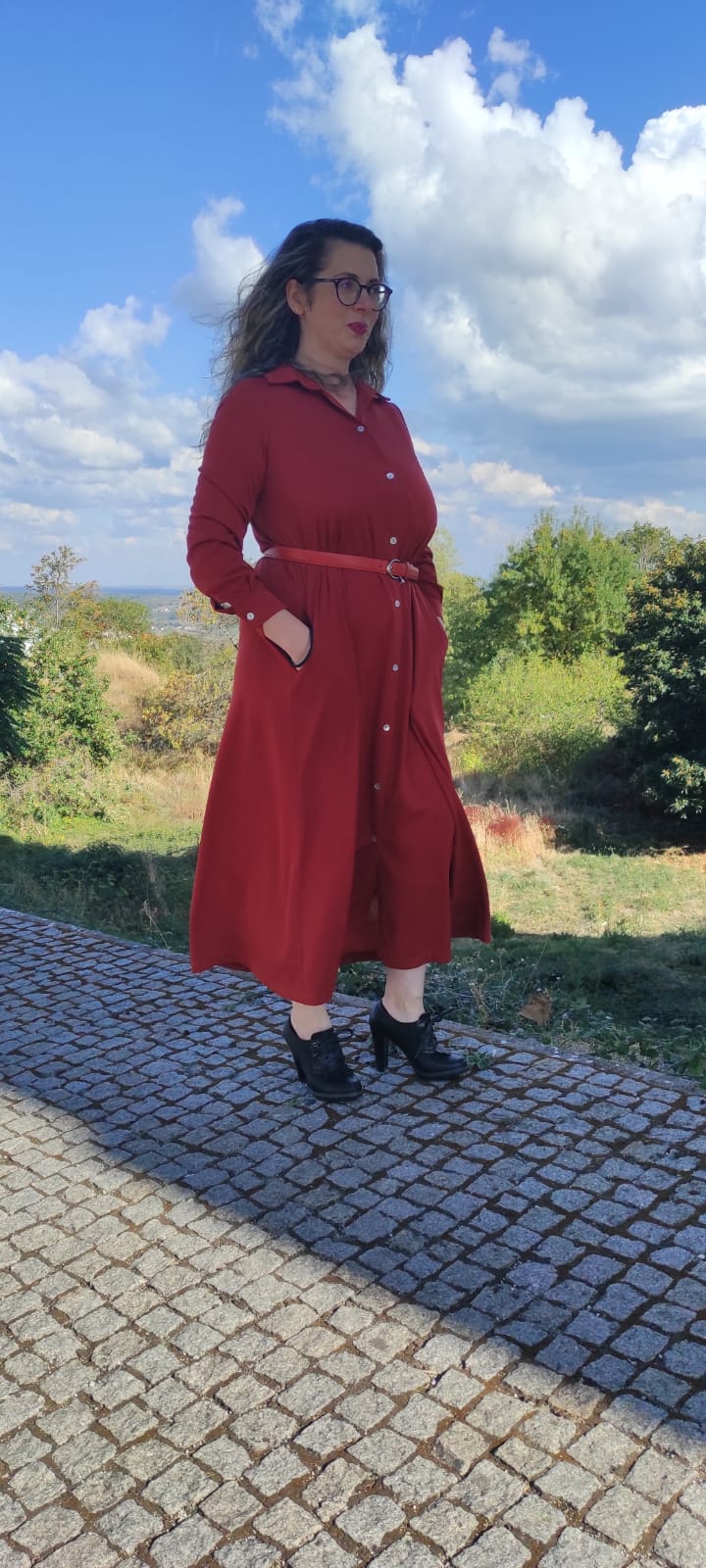 Mulher com vestido vermelho tijolo e sapatos pretos, ao ar livre em calçada de pedras e vegetação.