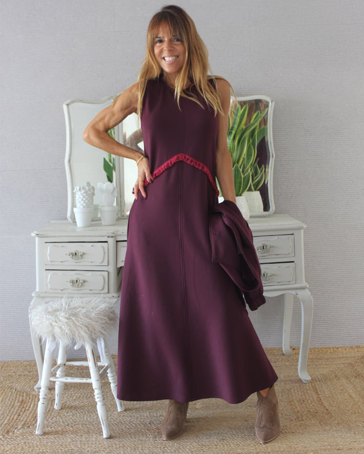 Mulher com vestido comprido bordeaux e botas castanhas em fundo interior com móvel branco e plantas