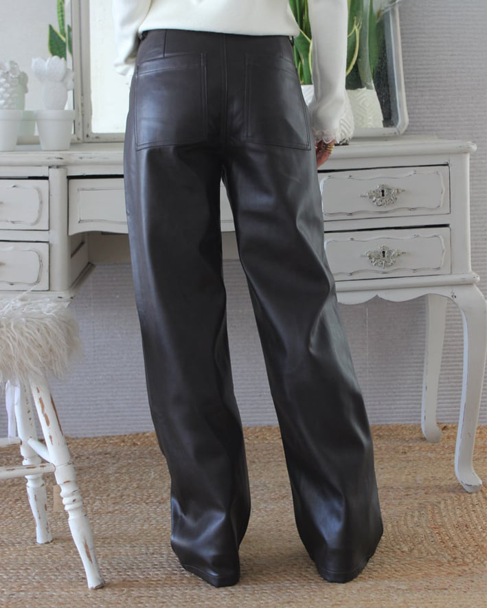 Calças de couro pretas largas com bolsos traseiros, pessoa vestindo blusa branca, móvel e cadeira brancos ao fundo