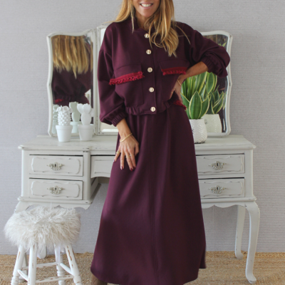 mulher com conjunto vinho de casaco com botões e saia comprida ao pé de móvel branco com planta e banco felpudo