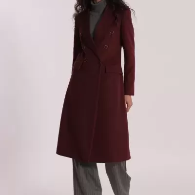 Mulher a usar casaco comprido bordô com botões, camisola cinzenta, calças cinzentas de riscas e sapatos pretos.