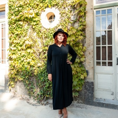 Mulher com vestido comprido preto e chapéu preto em ambiente exterior com parede coberta de hera e porta de vidro branca