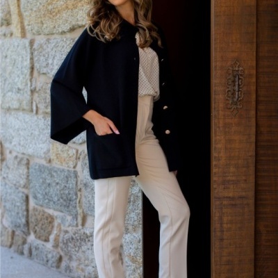 Mulher vestindo casaco preto, blusa branca com bolinhas, calças bege e botas bege junto a parede de pedra e porta de madeira