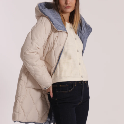 Mulher com casaco acolchoado bege e capuz azul cinzento, camisa creme e calças de ganga escuras.