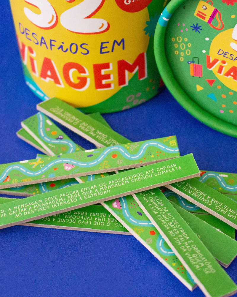 52 Desafios em Viagem Tiras de papel verdes com texto e ilustrações de estradas ao lado de um cilindro colorido com texto