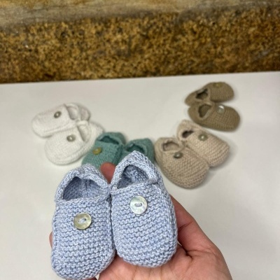 Cinco pares de sapatinhos de bebé em malha com botão em várias cores, sobre superfície branca, parede de pedra ao fundo