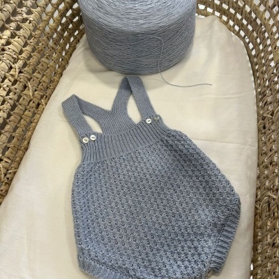Macacão de malha cinza claro com botões colocado em cesta de vime com novelo cinza.