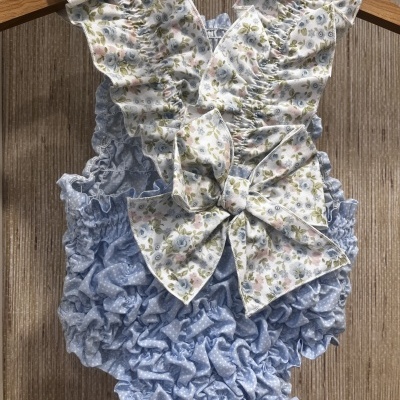 Macacão para bebé azul claro com padrão floral e laço em tecido, pendurado numa cabide de madeira