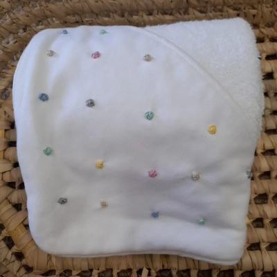 Toalha branca de bebé com capuz e pequenos bordados coloridos em pontos, sobre fundo de materiais naturais trançados
