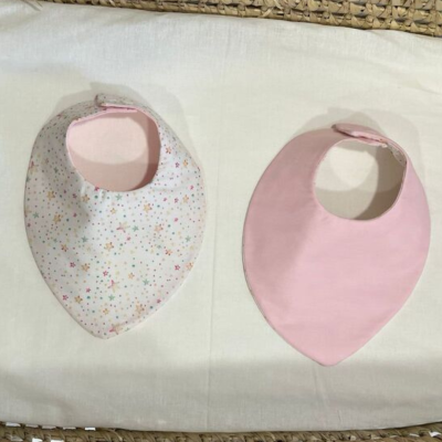 Conjunto de dois babetes de bebé, um branco com estrelas coloridas e outro rosa liso, em fundo branco com borda trançada.