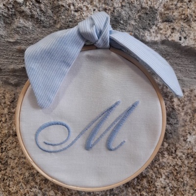 Bastidor redondo com letra bordada e fita azul claro amarrada no topo