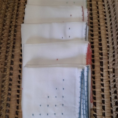 Seis panos de cozinha brancos com bordados coloridos empilhados sobre superfície de fibras naturais