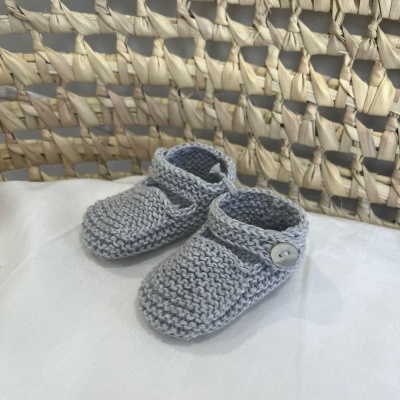 Sapatinhos de bebé em malha cinzenta com botão, sobre tecido branco, com cadeira de vime ao fundo