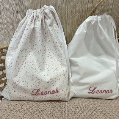 Dois sacos de pano brancos com nome Leonor, um com estrelas coloridas, sobre tecido bege e cesto de vime