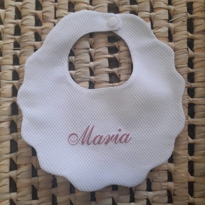 Babete branco com nome bordado e bordas onduladas sobre superfície de fibras entrelaçadas