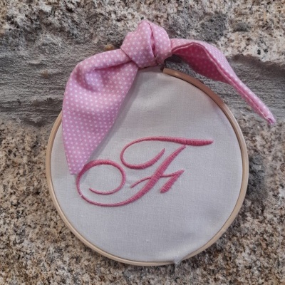 Bastidor com tecido branco bordado com letra F em vermelho e fita rosa de bolinhas sobre pedra rústica