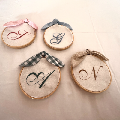 Quatro bastidores redondos de bordado com letras bordadas e fitas decorativas sobre fundo creme.