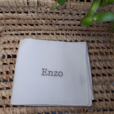 Guardanapo branco com bordado Enzo sobre tábua de palha e ramos verdes