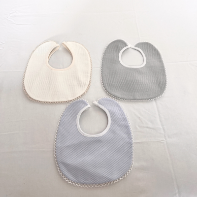 Três babetes de tecido em bege, cinza e azul claro com acabamento em renda, sobre superfície branca.