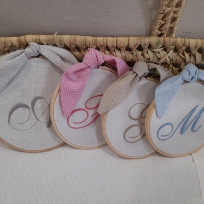 Quatro aros de bordado com letras bordadas e laços coloridos sobre superfície branca e cesta de palha