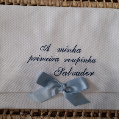 Fralda branca bordada com texto e laço azul claro sobre superfície trançada